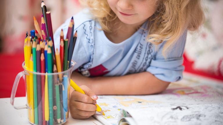  Blonde child colouring in with a jar full of colourful pencils.